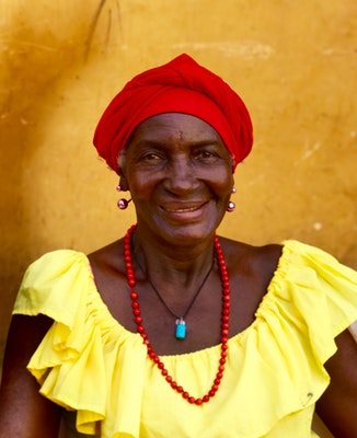 elder African woman with red headwrap, yellow ruffled dress, and red bead necklace