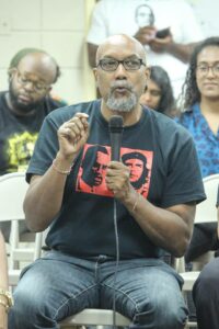 Ajamu Baraka in a black and red shirt, sitting with a mic, with people sitting behind.