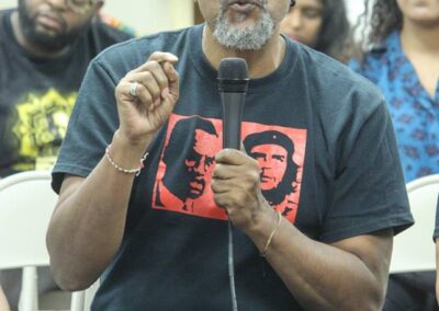 Ajamu Baraka in a black and red shirt, sitting with a mic, with people sitting behind.